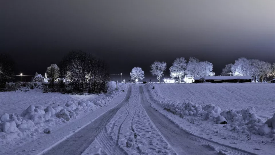 Mindre väg i snötäckt landskap efter mörkrets inbrott med en gård och träd vid horisonten. Foto