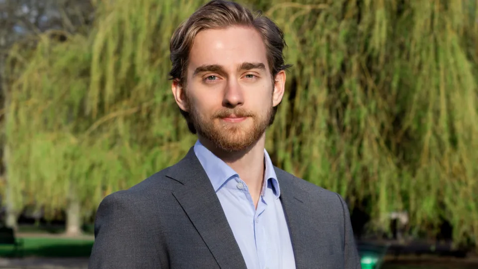 Foto på Daniel. Han tittar i kameran och har på sig en kostym. Bakgrunden är ljus och grön.