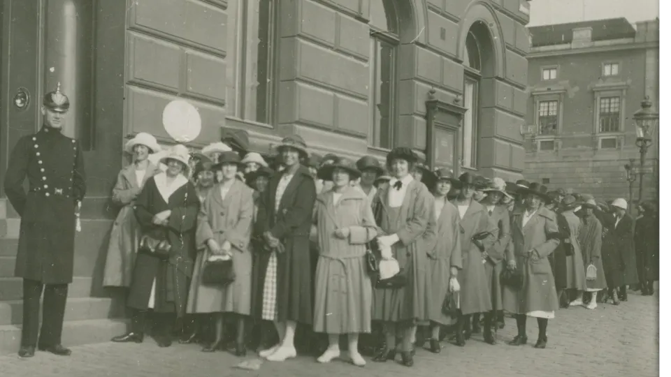 En polis och flera kvinnor utanför ett stort hus. Svartvitt foto.